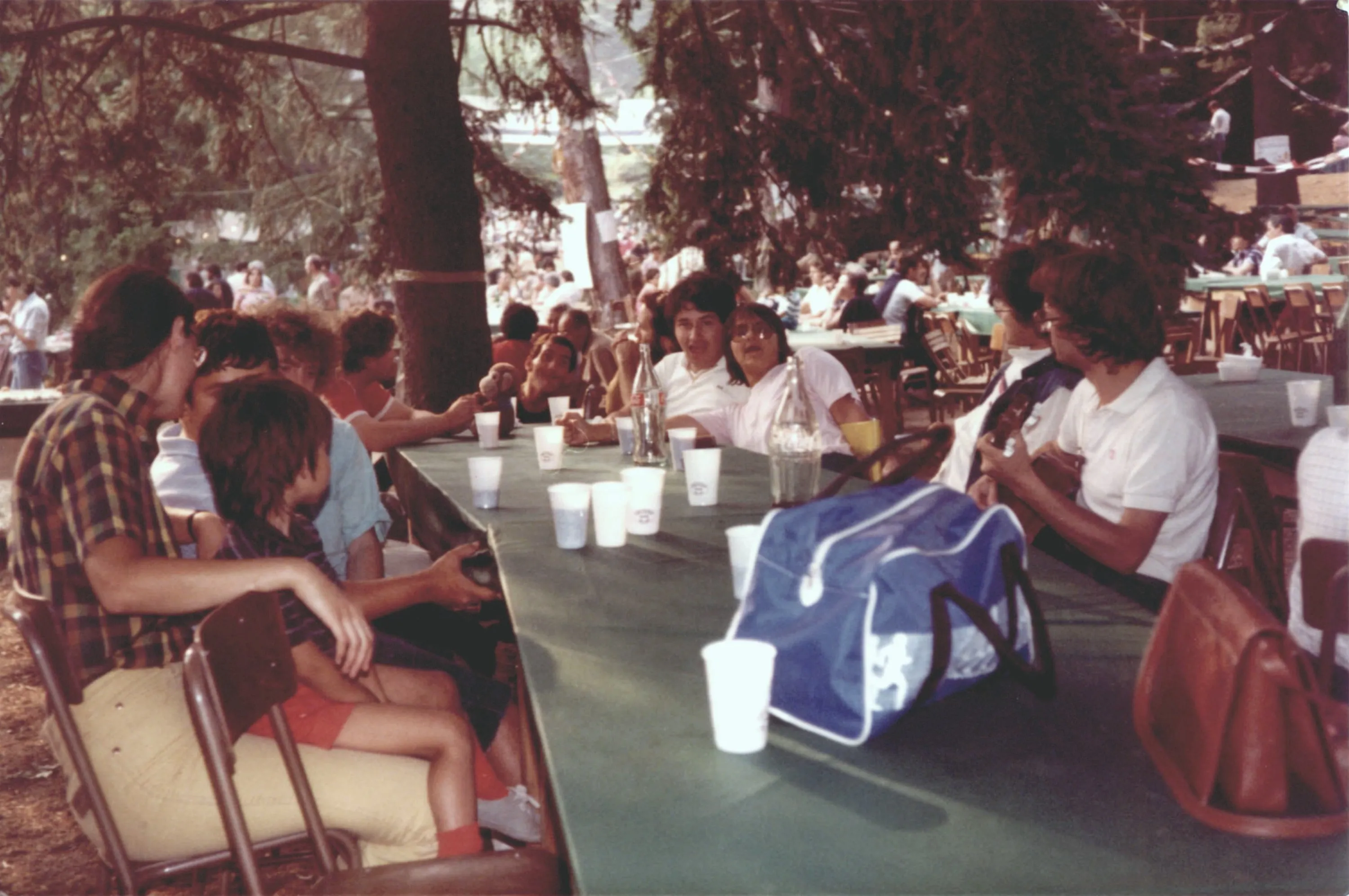Una fotografia d'epoca: ragazzi e volontari seduti a una lunga tavolata all'aperto, sotto gli alberi, durante una festa.