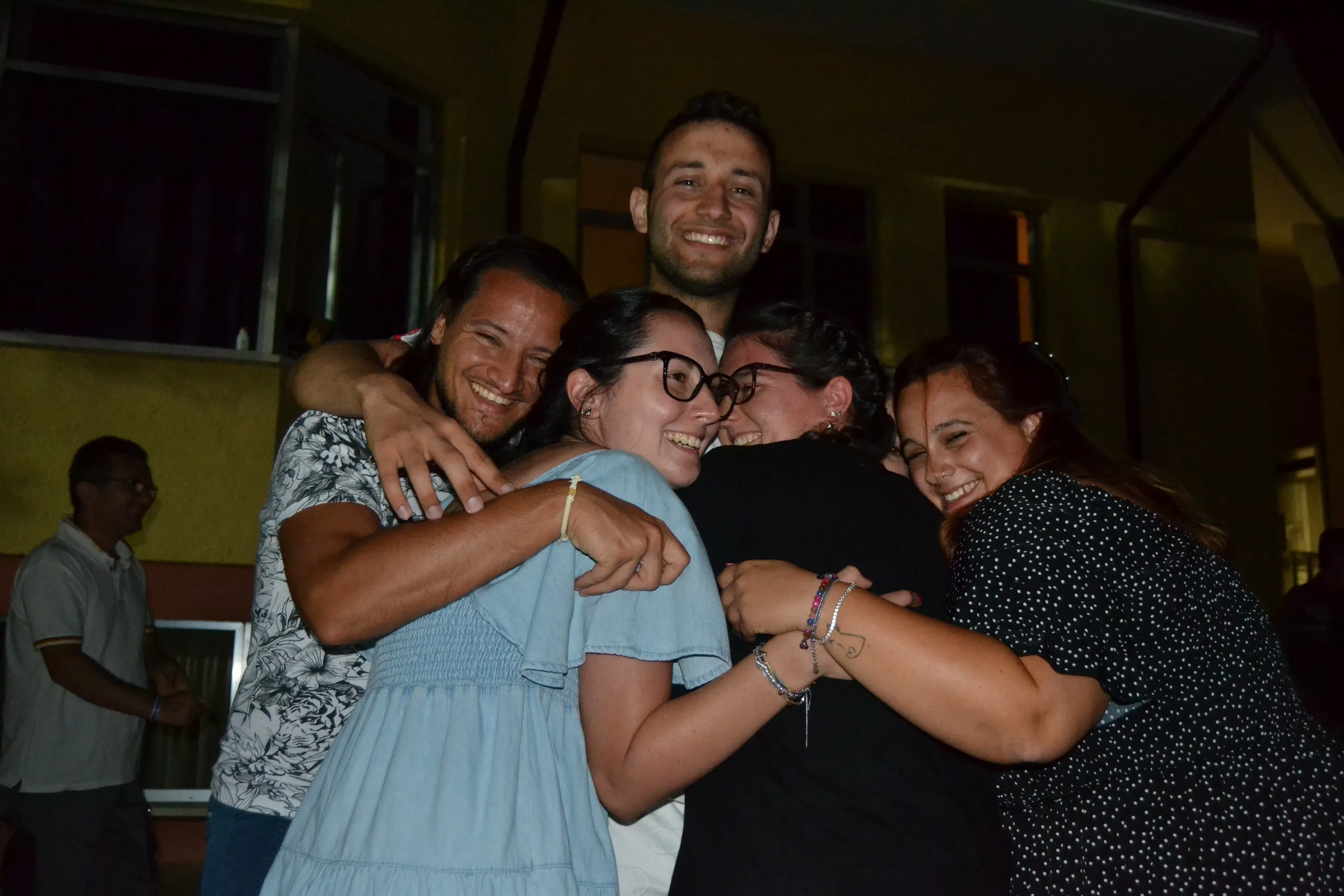 Cinque ragazzi abbracciati e sorridenti durante una festa all'aperto, di sera.